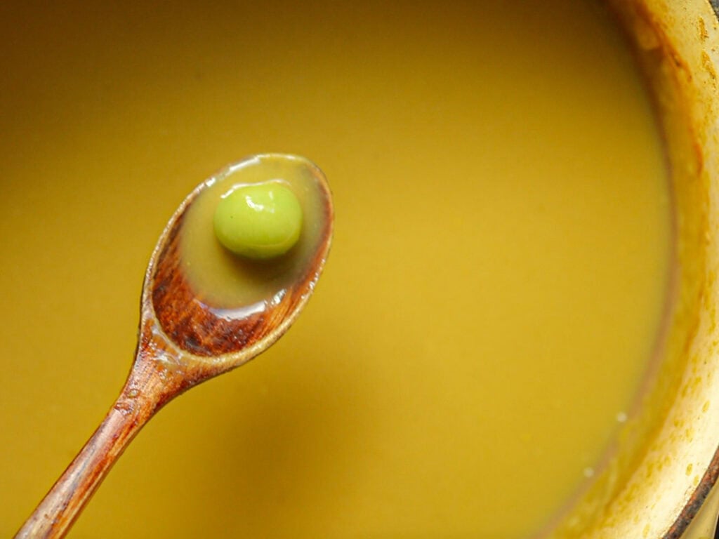Creamy Broccoli Soup with Dumplings - delicious and Gluten-Free! 17 A wooden spoon hovers over a bowl of smooth, creamy broccoli soup, holding a single green rice dumpling. The soups surface is reflective and looks rich and thick. The scene is close-up, focusing on the contrasting textures and colors.