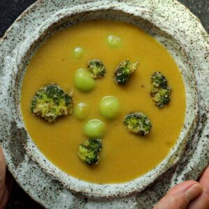 Creamy Broccoli Soup with Dumplings - delicious and Gluten-Free! 21 A rustic ceramic bowl brimming with creamy broccoli soup, adorned with roasted florets and green dumplings. Hands gently cradle the bowl, emphasizing the earthy texture of the pottery.