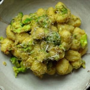 Easy Creamy Broccoli Gnocchi - Healthy and Delicious. 22 A bowl of creamy broccoli gnocchi mixed with chopped broccoli and topped with grated cheese, all coated in a light greenish-brown sauce. The dish is served in a rustic, light-colored ceramic bowl.