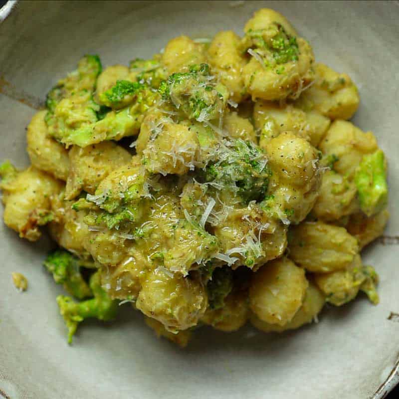 30-Minute Delicious Asparagus Pasta Recipe (Vegan). 22 A bowl of creamy broccoli gnocchi mixed with chopped broccoli and topped with grated cheese, all coated in a light greenish-brown sauce. The dish is served in a rustic, light-colored ceramic bowl.
