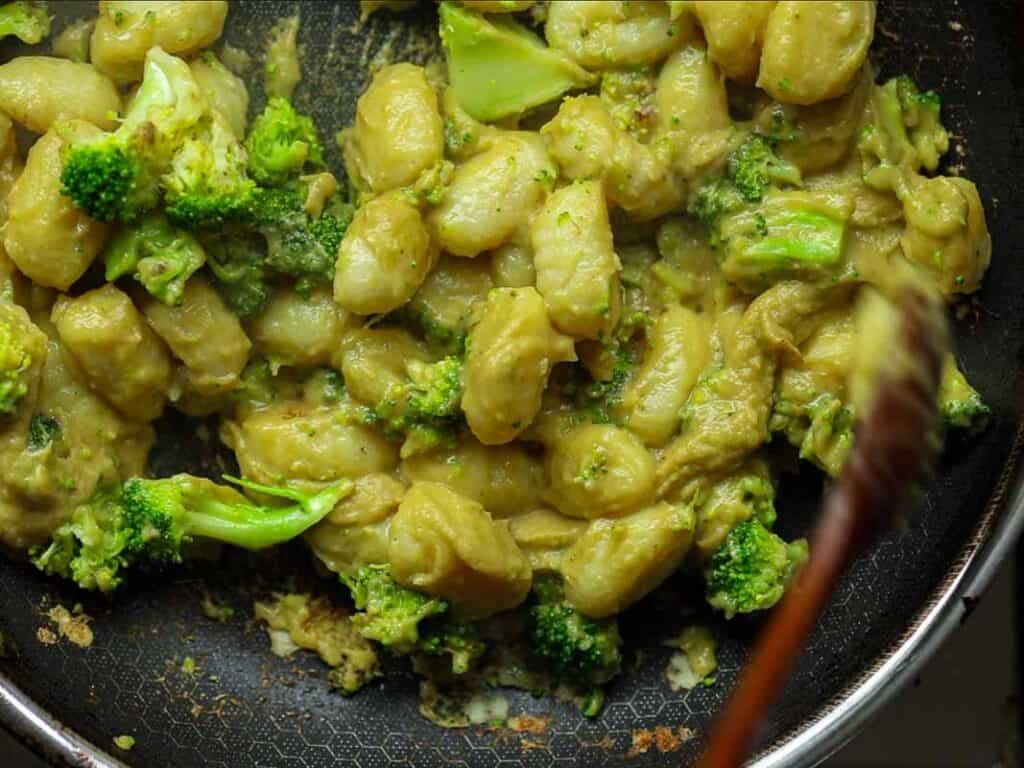 Easy Creamy Broccoli Gnocchi - Healthy and Delicious. 2 A frying pan filled with creamy gnocchi and broccoli florets, stirred with a wooden spoon. The gnocchi is coated in a thick, light brown sauce. The dish appears savory and freshly cooked.