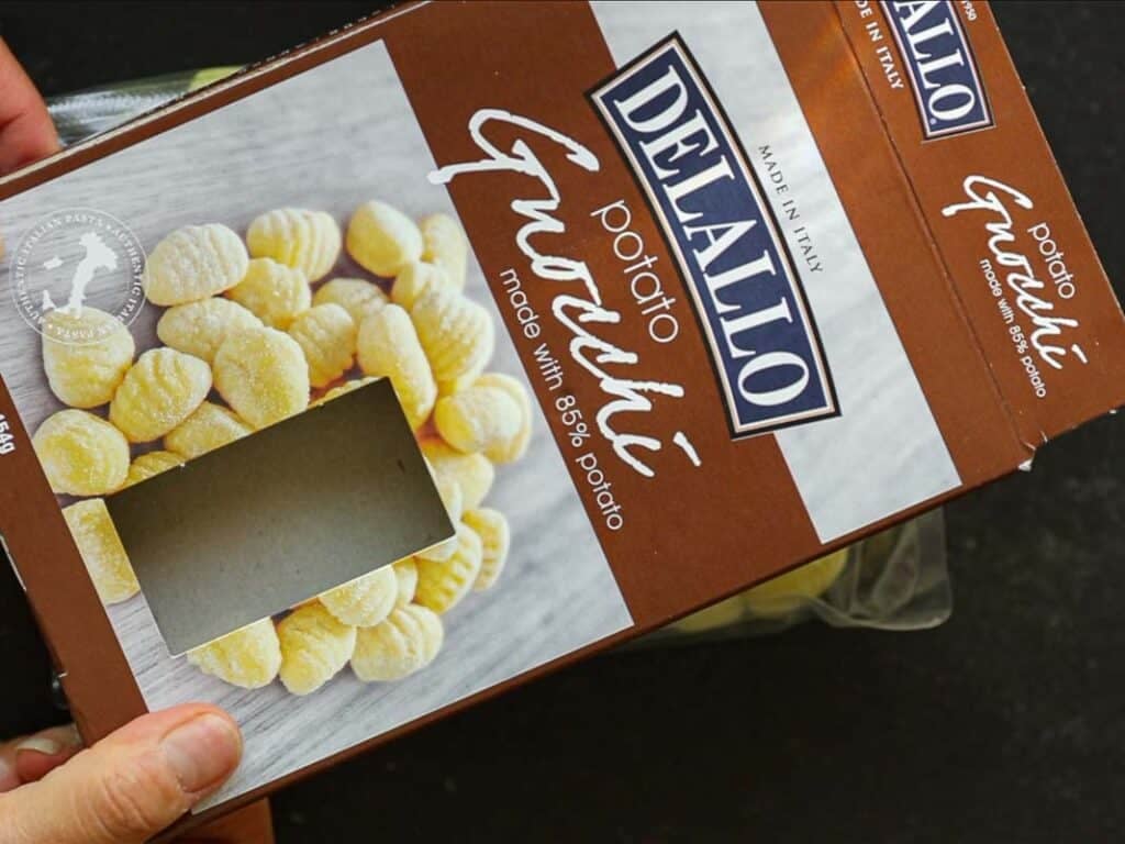 Easy Creamy Broccoli Gnocchi - Healthy and Delicious. 6 A person holds a brown DeLallo Potato Gnocchi box with a clear window showing gnocchi inside. The box text reads Made with 85% potato. The box is placed on a dark surface.