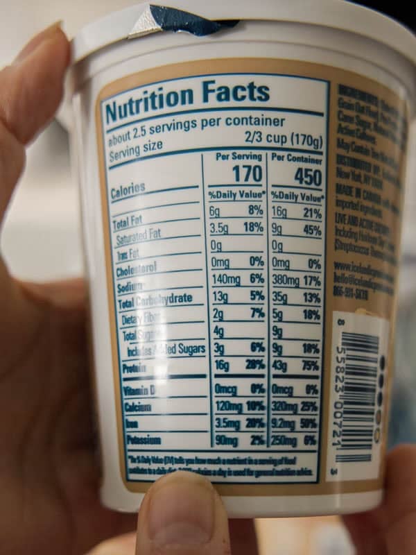 Easy & Delicious 3-INGREDIENT Bagels (vegan and gf option). 6 Close-up of a hand holding a container, focusing on the nutrition facts label. It lists calories, fats, cholesterol, sodium, carbohydrates, protein, and vitamins, with values per serving and per container. A thumb is visible at the bottom.