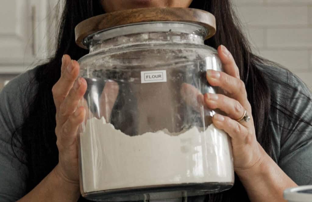 Easy & Delicious 3-INGREDIENT Bagels (vegan and gf option). 4 A person holding a large glass jar labeled Flour with a wooden lid, partially filled with flour. The person is wearing grey long sleeves and has long dark hair, standing in a kitchen with a tiled backsplash in the background.