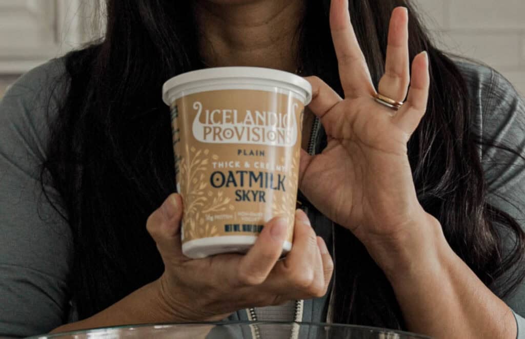 Easy & Delicious 3-INGREDIENT Bagels (vegan and gf option). 5 A person with long dark hair holds a container of Icelandic Provisions plain oatmilk skyr. Their left hand is making an OK gesture, and they are wearing a gold ring.