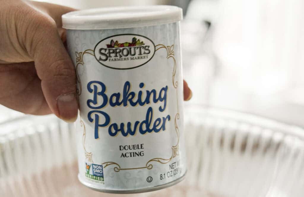 Easy & Delicious 3-INGREDIENT Bagels (vegan and gf option). 7 A hand holds a can of Sprouts Farmers Market Baking Powder over a metal baking pan. The label indicates it is double acting and non-GMO verified. The can has a white lid and decorative design.