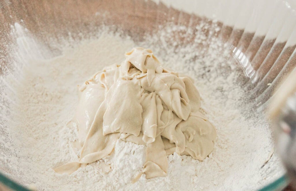 Easy & Delicious 3-INGREDIENT Bagels (vegan and gf option). 8 A large glass bowl contains a mound of creamy hi-protein yogurt placed on a bed of white flour. The mixture appears smooth and ready for kneading or further preparation. The transparent bowl shows a hint of a wooden surface underneath.