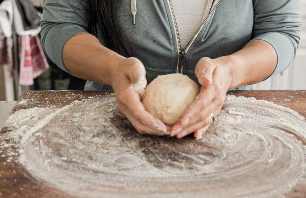 Easy & Delicious 3-INGREDIENT Bagels (vegan and gf option). 9 Person shaping a ball of dough with their hands on a floured wooden surface. They are wearing a grey zip-up hoodie, and the kitchen background is slightly blurred.