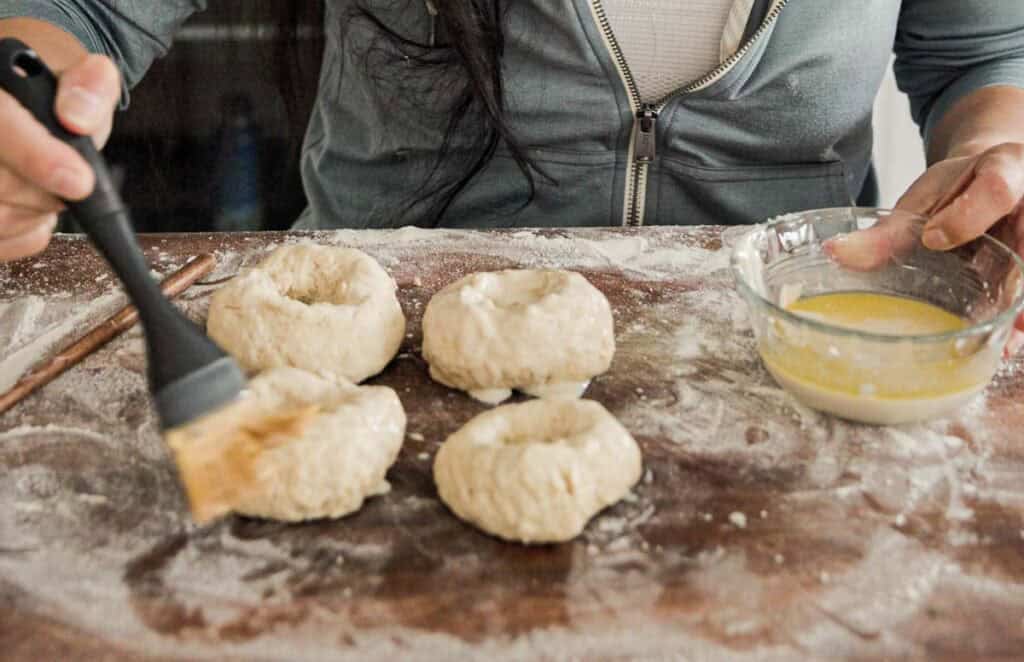 Easy & Delicious 3-INGREDIENT Bagels (vegan and gf option). 16 Person brushing wash onto four raw bagels on a floured wooden surface. A glass bowl with egg wash is nearby. The person is wearing a zip-up hoodie.