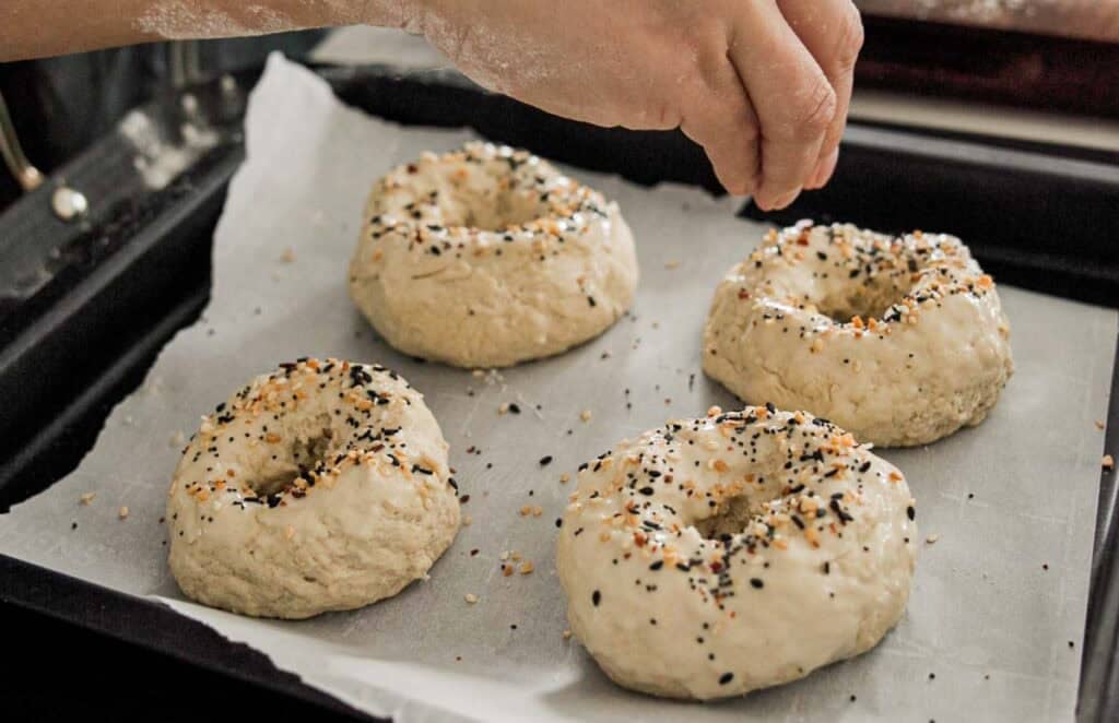 Easy & Delicious 3-INGREDIENT Bagels (vegan and gf option). 17 A persons hand sprinkles seasoning onto four uncooked bagels resting on a parchment-lined baking sheet. The bagels are topped with a mix of black and white seeds. Flour dust is visible on the fingers.