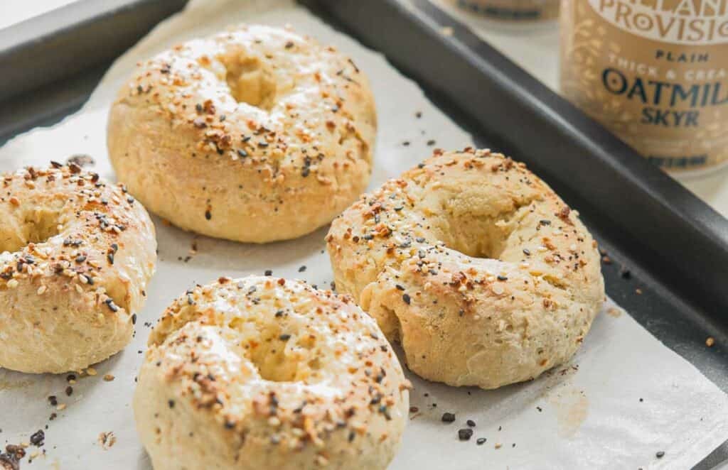 Easy & Delicious 3-INGREDIENT Bagels (vegan and gf option). 18 Four freshly baked bagels with seasoning on top are placed on a parchment-lined baking tray. A container of oatmilk skyr is visible in the background.