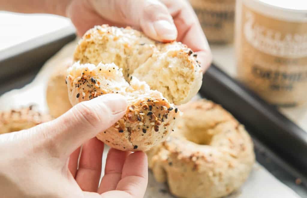 Easy & Delicious 3-INGREDIENT Bagels (vegan and gf option). 2 Close-up of hands breaking apart a freshly baked 3-ingredient bagel topped with seeds, revealing a fluffy texture inside. Other bagels are visible on a parchment-lined tray in the background.