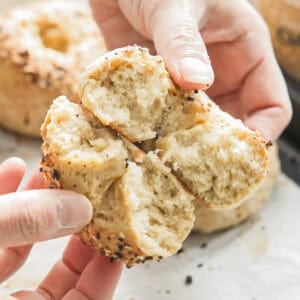 Easy & Delicious 3-INGREDIENT Bagels (vegan and gf option). 21 Hands breaking apart a freshly baked 3-ingredient bagel topped with seeds. The interior appears soft and fluffy, while the crust is golden brown and slightly crispy, with visible poppy and sesame seeds scattered on the surface.