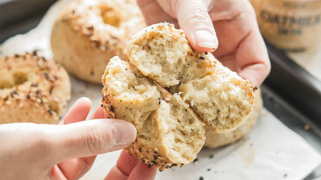 Easy & Delicious 3-INGREDIENT Bagels (vegan and gf option). 1 Hands tearing apart a freshly baked, sesame seed-crusted 3-ingredient bagels, revealing a soft, fluffy interior. Other bagels on a parchment-lined tray are visible in the background.