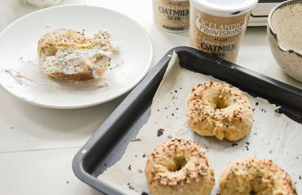 Easy & Delicious 3-INGREDIENT Bagels (vegan and gf option). 3 Freshly baked bagels on a baking sheet with one bagel cut and spread with a creamy topping on a plate. Containers of Icelandic Provisions plain oat milk skyr are in the background on a light-colored surface.