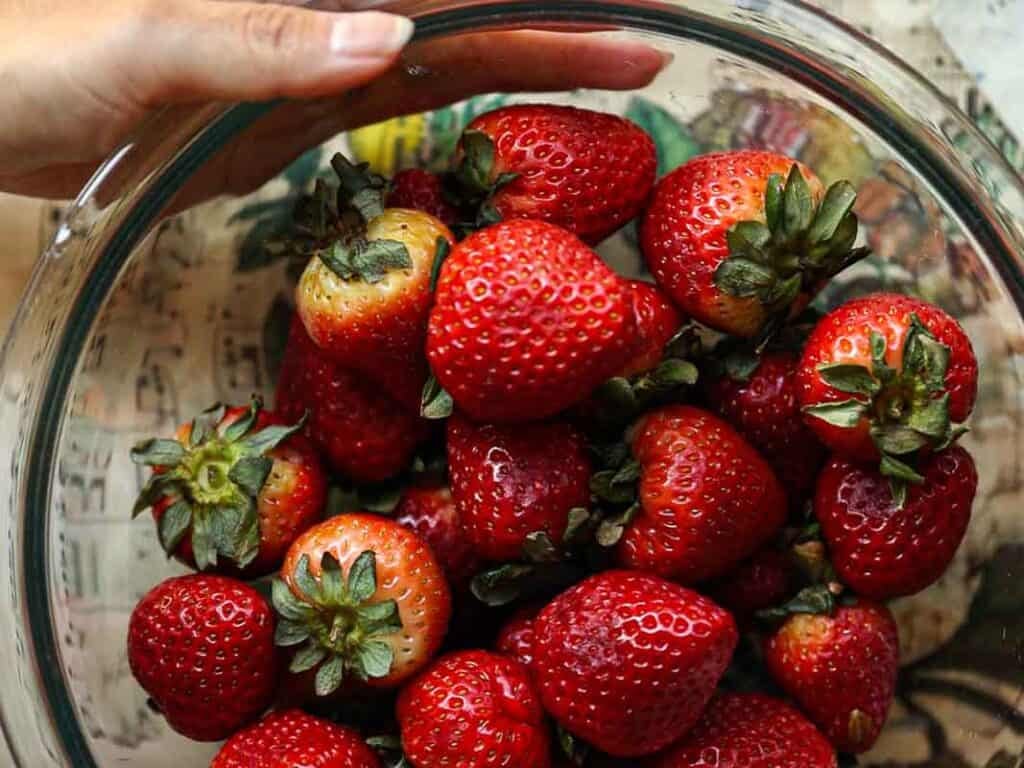 Stunning Pink Vegan Strawberry Cake (Fresh berries). 4 A hand holding a glass bowl filled with bright red strawberries, some with leaves still attached. The background features a colorful, patterned surface.