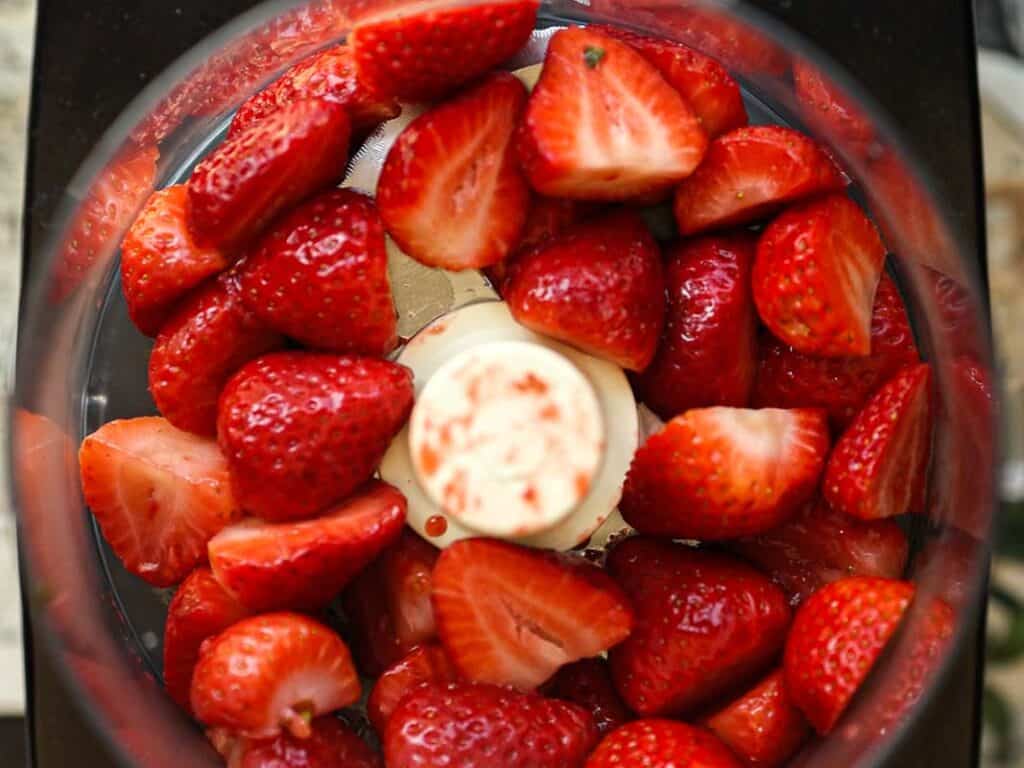 Stunning Pink Vegan Strawberry Cake (Fresh berries). 9 Top view of halved strawberries arranged in a circular pattern inside a food processor, with the cream-colored center of the processor visible. The strawberries are bright red and glossy, contrasting with the dark outer rim of the processor.