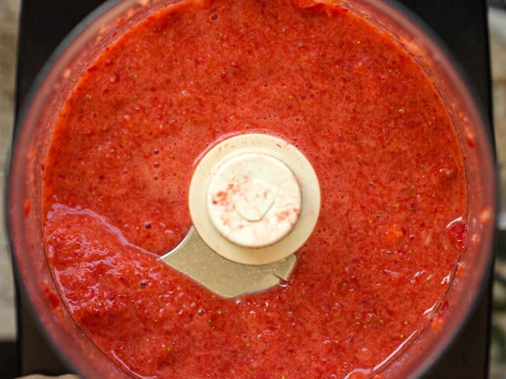 Stunning Pink Vegan Strawberry Cake (Fresh berries). 10 Top view of a food processor containing a smooth, bright red mix of blended strawberries. The processors blade is visible, partially covered by the fruit puree. The background is a blurred countertop.