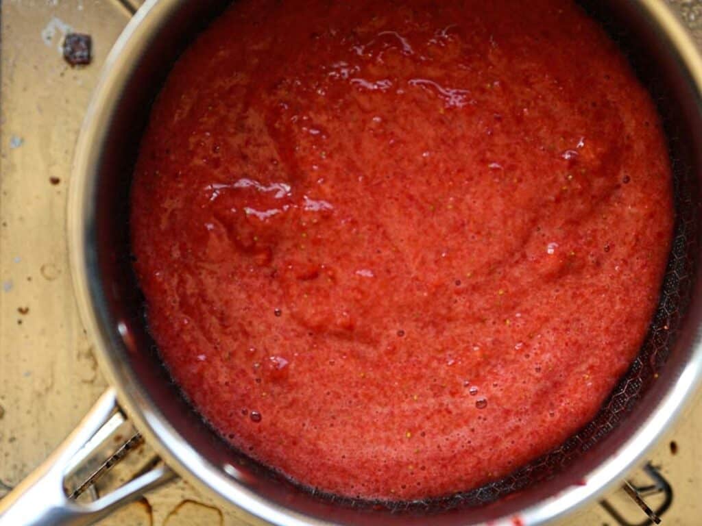 Stunning Pink Vegan Strawberry Cake (Fresh berries). 11 A close-up of a pot filled with a smooth red sauce, likely tomato or berry-based, placed on a beige countertop. The sauce appears thick and evenly mixed, with a glossy texture.