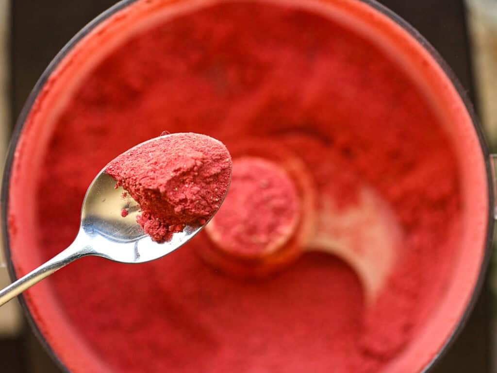 Stunning Pink Vegan Strawberry Cake (Fresh berries). 14 A close-up of a spoon holding bright red powder, hovering above a food processor filled with more of the same substance. The processors metal blade is visible amidst the vibrant powder.