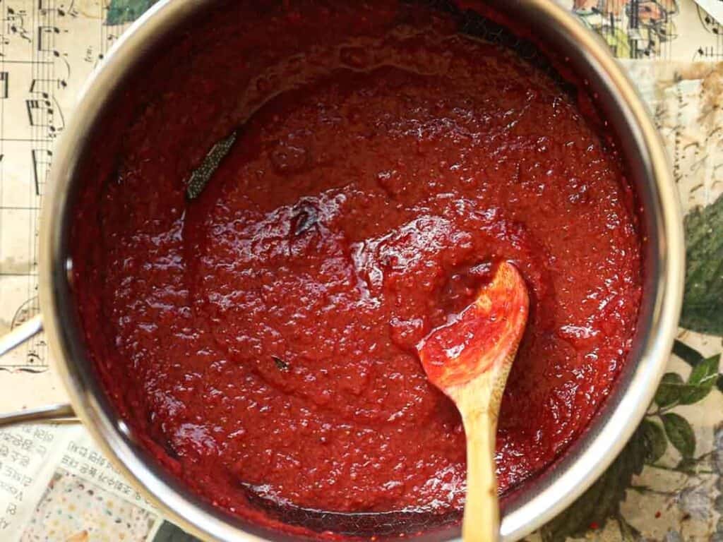 Stunning Pink Vegan Strawberry Cake (Fresh berries). 12 A pot filled with rich red strawberry reduction sauce, with a wooden spoon partially submerged. The sauce has a thick, smooth texture. The pot rests on a surface covered with printed paper or newspaper.