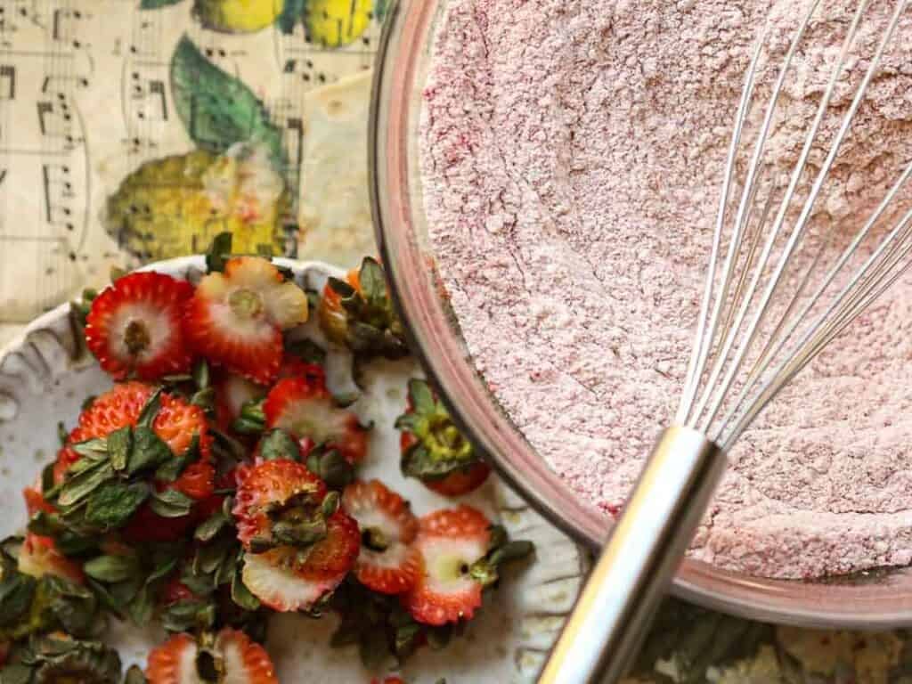Stunning Pink Vegan Strawberry Cake (Fresh berries). 15 A close-up view of a mixing bowl filled with pinkish flour and a whisk. Beside the bowl, there are sliced strawberries with green tops on a plate. The background features a decorative music note pattern with yellow accents.