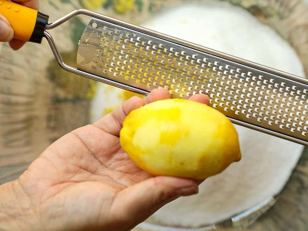 Stunning Pink Vegan Strawberry Cake (Fresh berries). 7 Close-up of hands holding a lemon and a zester with yellow handle. Lemon peel is partially grated. A white bowl is visible underneath, catching the lemon zest.