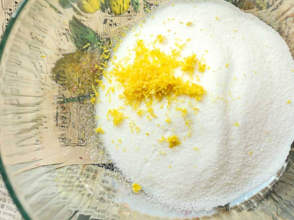 Stunning Pink Vegan Strawberry Cake (Fresh berries). 17 A clear glass bowl contains a mixture of white granulated sugar and finely grated lemon zest. The contrast between the vibrant yellow zest and the white sugar is highlighted against a newspaper background.