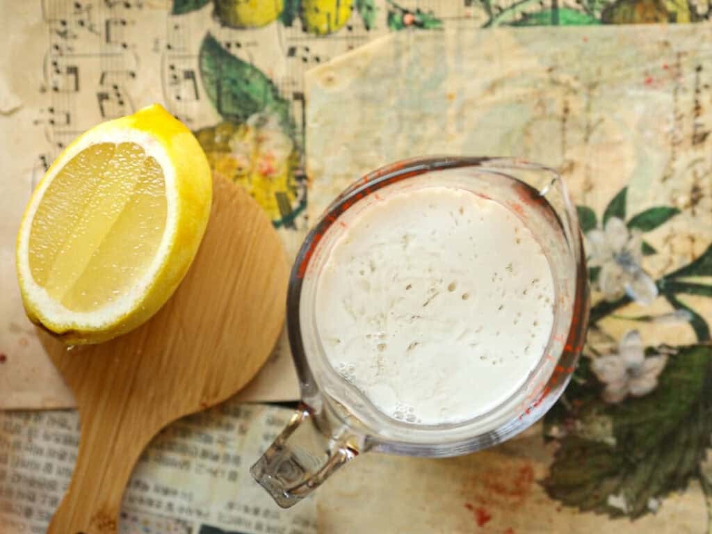 Stunning Pink Vegan Strawberry Cake (Fresh berries). 8 A sliced lemon on a wooden board next to a glass measuring cup filled with a frothy liquid. The background features a botanical pattern with flowers and out-of-focus sheet music.