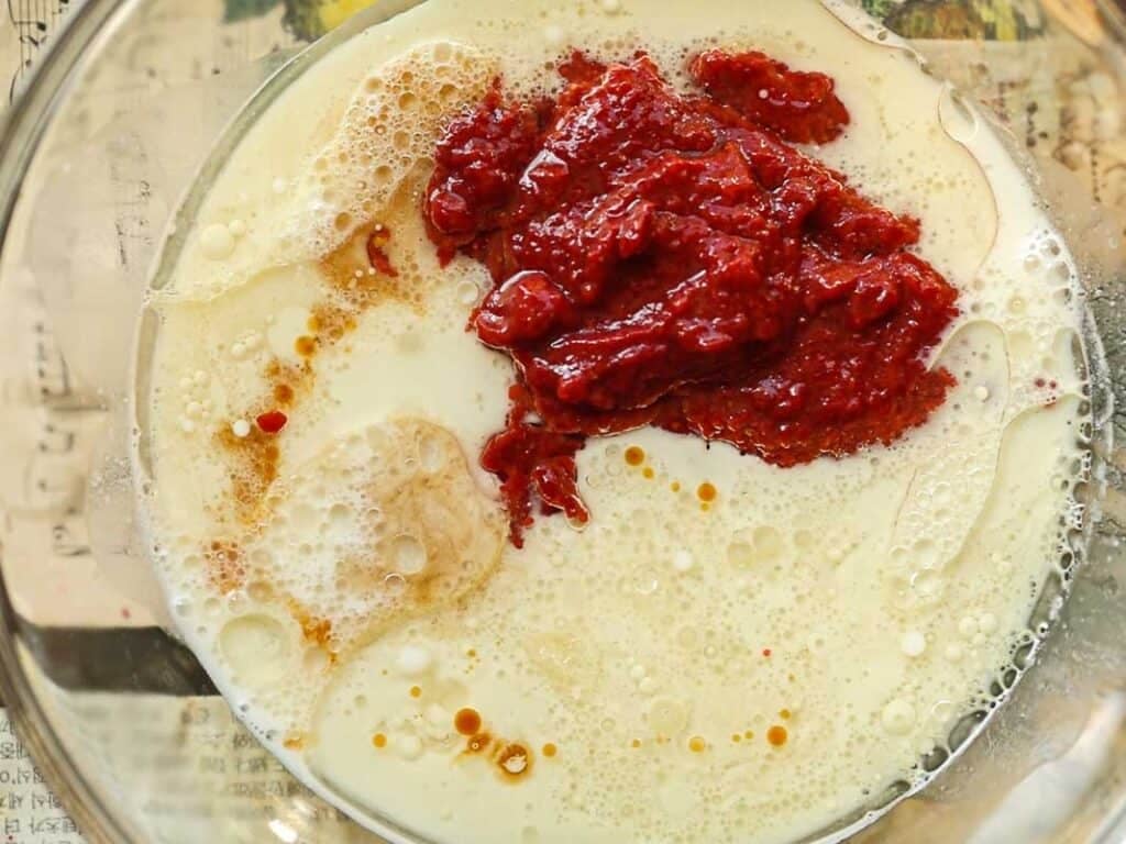 Stunning Pink Vegan Strawberry Cake (Fresh berries). 20 A glass bowl containing milk, strawberry reduction, and other ingredients for strawberry cake batter. The ingredients are partially mixed, with some floating bubbles visible. The bowl is placed on a beige surface with faint text.