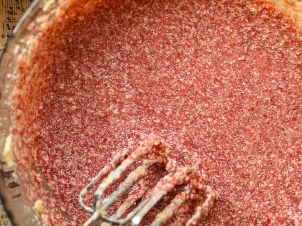 Close-up view of a mixing bowl filled with a pink, speckled batter. An electric mixer with pink-tinted beaters is partially submerged, indicating the batter has been recently mixed. The background shows part of a music sheet.