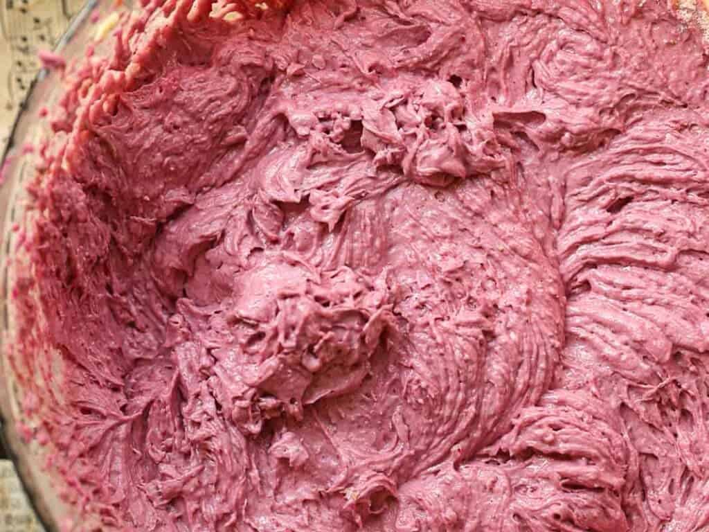 Close-up of a bowl filled with a thick, vividly purple batter. The texture is creamy and slightly lumpy, with swirls indicating it has been recently stirred. The background is partially visible, showing a pattern of text.