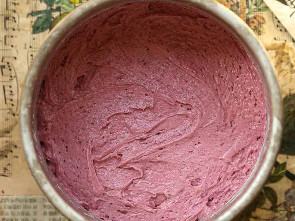A round cake pan filled with creamy, smooth purple cake batter. The batter is swirled and sits on a surface covered with sheet music.