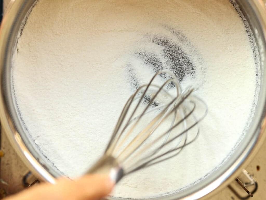 A hand holds a whisk, mixing flour and sugar in a small pot. The flour is evenly spread, with some traces of whisking visible. The scene suggests preparation for baking or cooking.