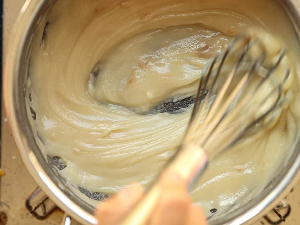 A hand whisking roux for ermine frosting in a stainless steel pot. The sauce has a smooth texture, and the whisk is partially submerged in the mixture. The pot is on a stovetop.