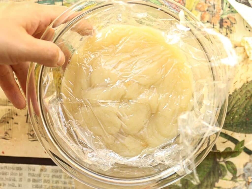 A hand holding a glass bowl covered with plastic wrap. Inside the bowl is roux for ermine frosting. The background features a colorful, printed tablecloth.