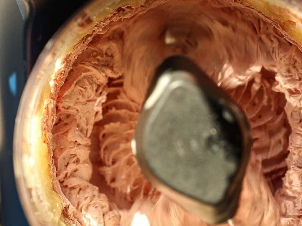 Close-up view of a food processor mixing creamy strawberry frosting. The rich, swirled texture is visible around the central blade, indicating a smooth consistency. The sides of the processor bowl show some chocolate residue.