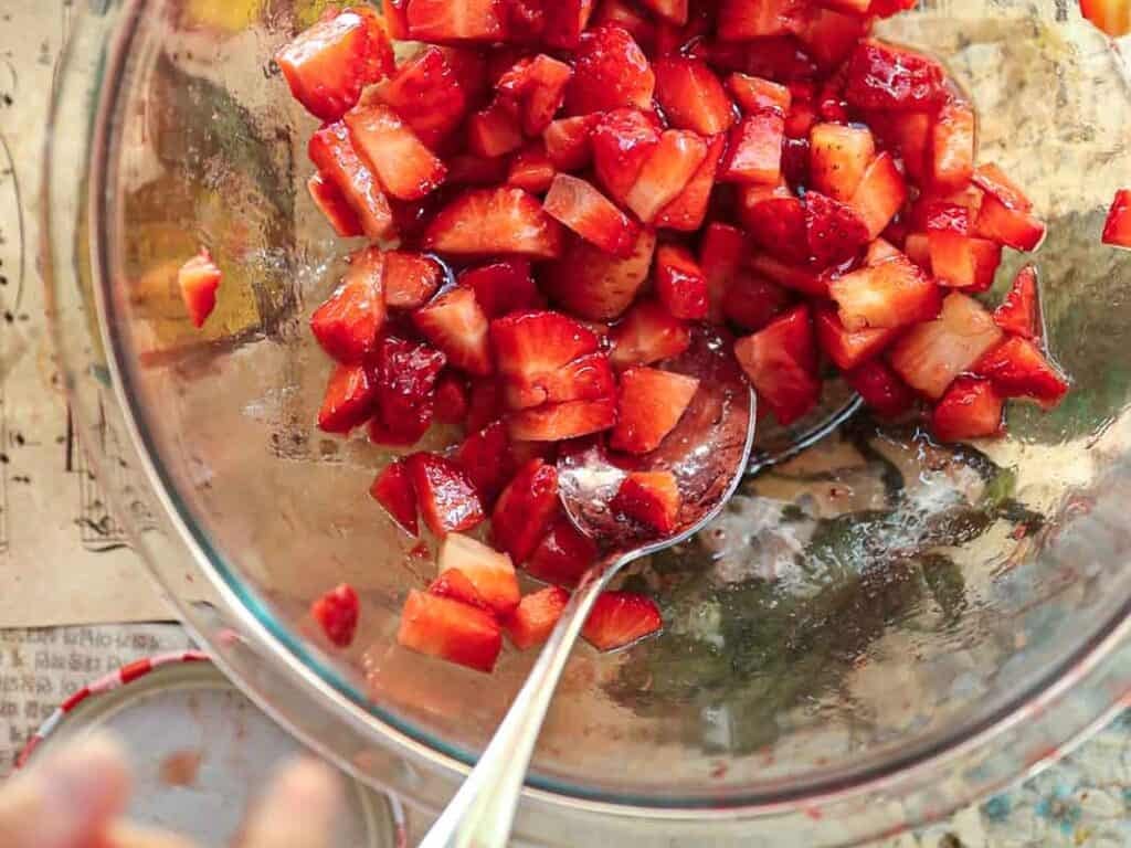 Stunning Pink Vegan Strawberry Cake (Fresh berries). 29 A glass bowl filled with diced strawberries. A silver spoon is partially submerged in the fruit mix. The background includes a tabletop with a decorative paper and a jar lid next to the bowl.