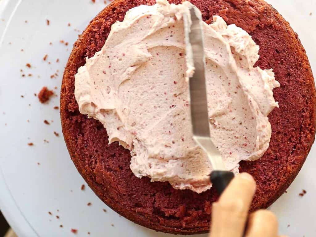 A person using an offset spatula to spread creamy frosting on a round vegan strawberry cake. The cake sits on a white surface, with some crumbs scattered around.