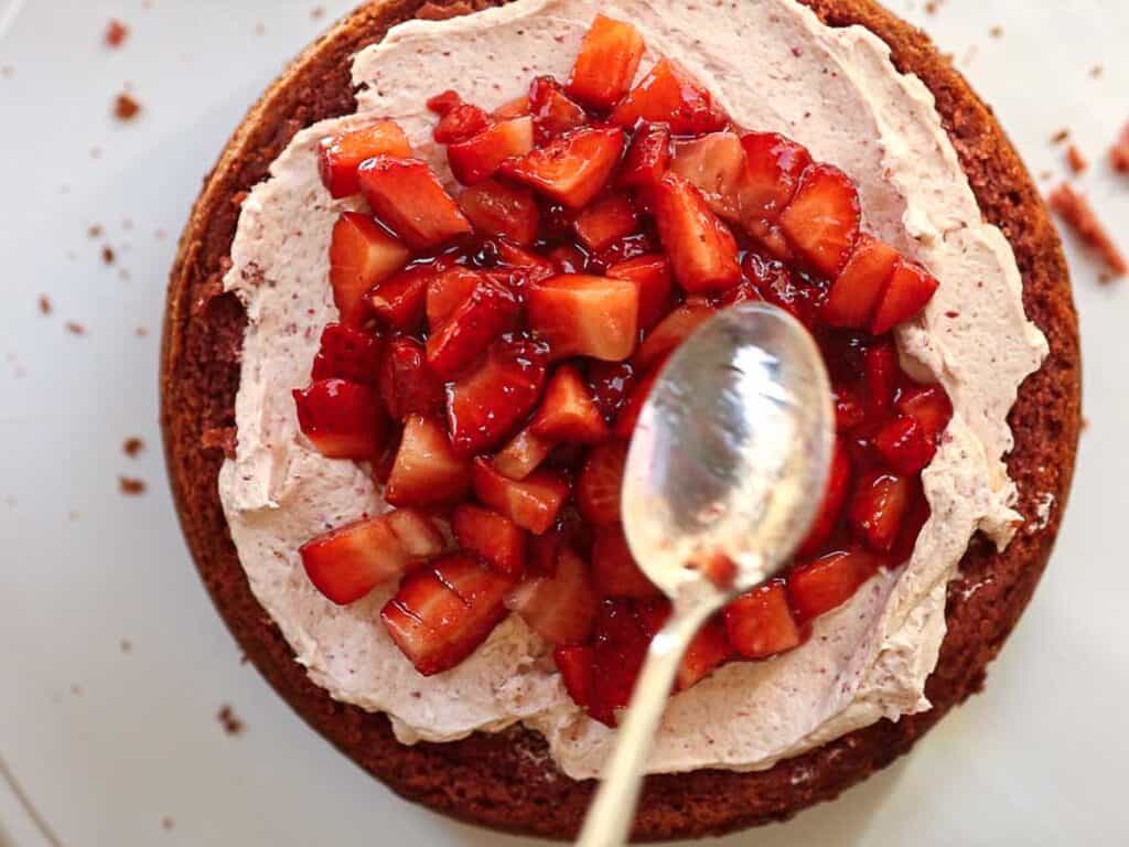 A round vegan strawberry cake with a layer of frosting is topped with chopped strawberries. A spoon is resting on top of the strawberries, suggesting the topping is being spread or adjusted. The cake is on a white surface with scattered crumbs around it.