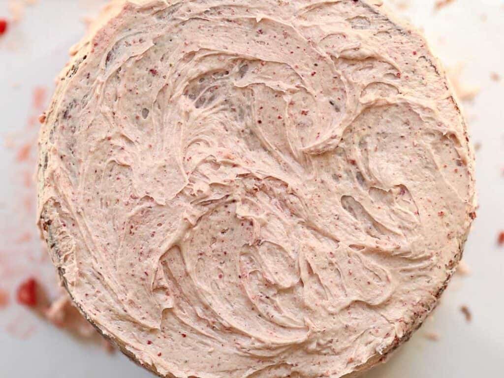 Stunning Pink Vegan Strawberry Cake (Fresh berries). 33 A close-up of a vegan strawberry cake topped with creamy, swirled frosting. The frosting has a light pink hue with small red specks, giving it a textured appearance. The cakes round shape is centered in the image.