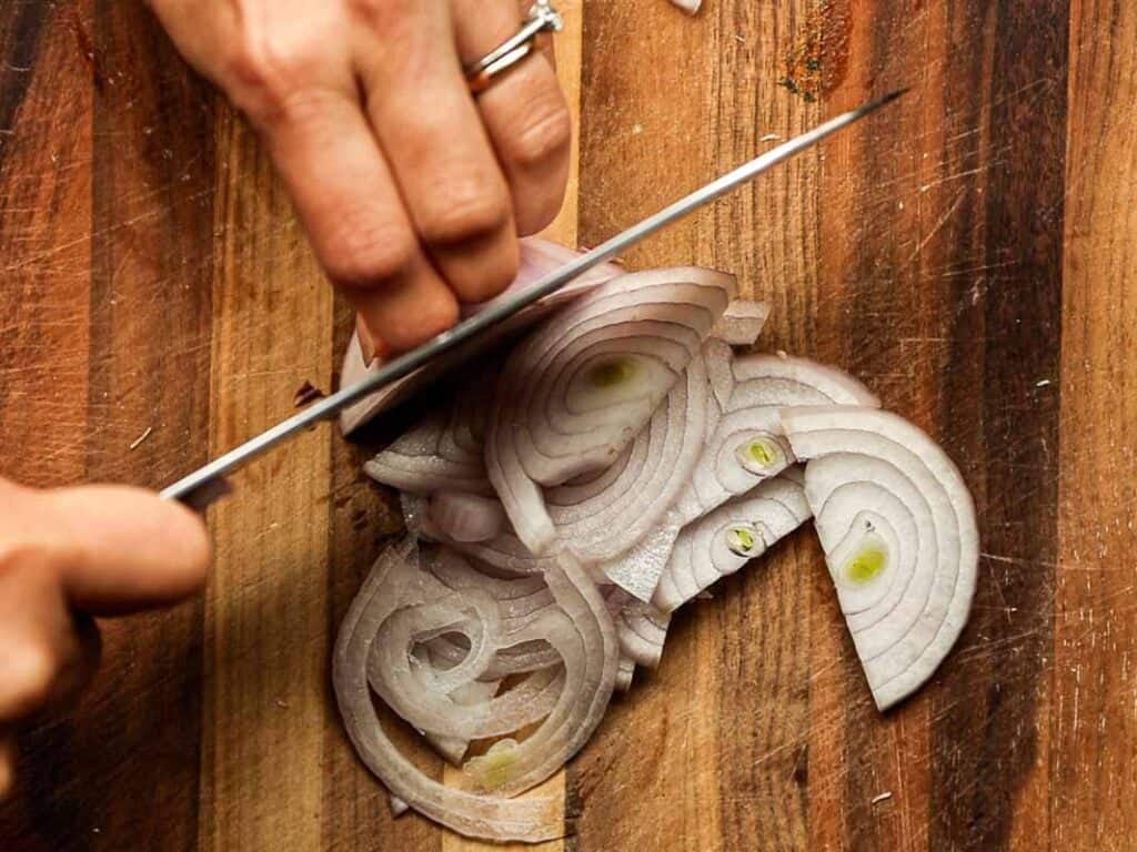 30-Minute Creamy Gochujang Vodka Pasta - Delicious & Dairy-FREE. 8 A close-up of a person slicing a shallot on a wooden cutting board. The shallot is cut into thin, circular slices. The person is wearing a ring and holding a knife with one hand, pressing down on the shallot with the other.