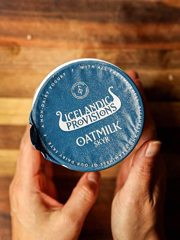 30-Minute Creamy Gochujang Vodka Pasta - Delicious & Dairy-FREE. 7 Close-up of hands holding a container with a blue lid labeled Icelandic Provisions Oatmilk Skyr. The lid is slightly pulled back on one side. The background is a wooden surface.