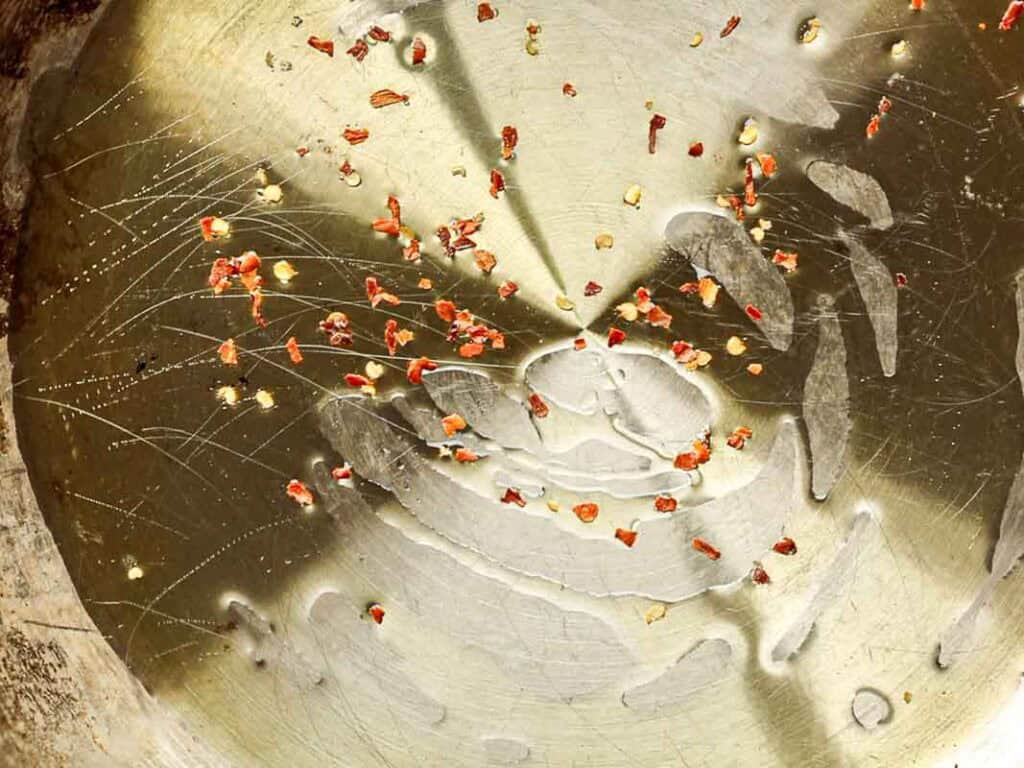 30-Minute Creamy Gochujang Vodka Pasta - Delicious & Dairy-FREE. 10 A close-up of a scratched metal surface with scattered red chili flakes and oil droplets, creating a textured and reflective appearance.