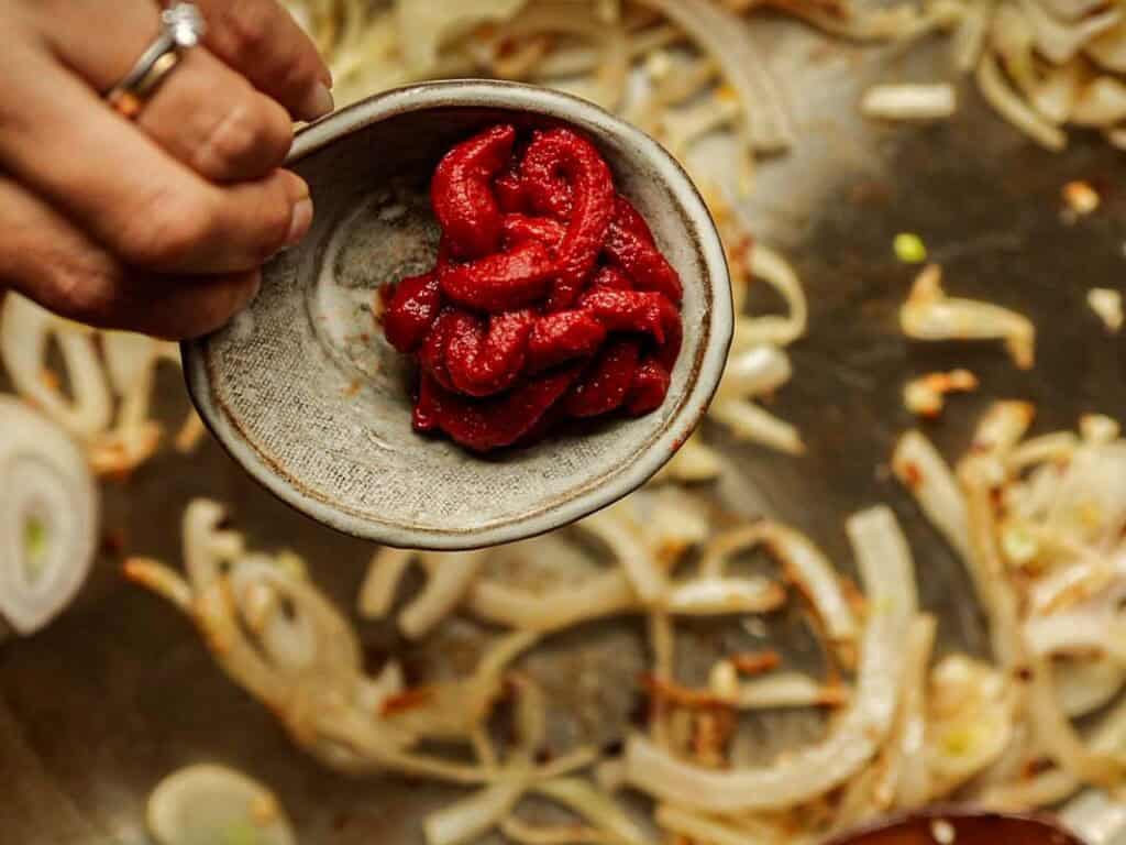 30-Minute Creamy Gochujang Vodka Pasta - Delicious & Dairy-FREE. 5 A hand holding a small ceramic bowl filled with red tomato paste, positioned above a pan with sautéing onions. The onions are browning and slightly translucent. The hand is adorned with rings on the fingers.