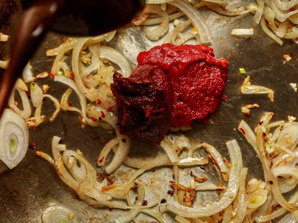 30-Minute Creamy Gochujang Vodka Pasta - Delicious & Dairy-FREE. 15 Onions and tomatoes are being cooked in a pan. The shallots are golden brown, and the tomatoes appear as a rich, deep red paste (gochujang and tomato paste), suggesting the beginnings of a sauce or stew.