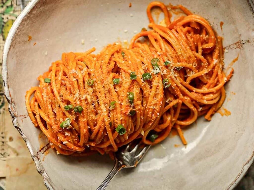 30-Minute Creamy Gochujang Vodka Pasta - Delicious & Dairy-FREE. 2 A bowl of vegan gochujang vodka pasta tossed in a rich red tomato sauce, garnished with green herbs and grated cheese. A fork rests beside the pasta, ready to twirl the strands. The dish is served in a rustic, shallow ceramic bowl.