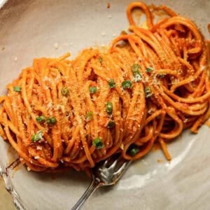 30-Minute Creamy Gochujang Vodka Pasta - Delicious & Dairy-FREE. 30 A close-up of a bowl of vegan gochujang vodka pasta coated in a rich red sauce, garnished with grated cheese and chopped green herbs. A fork rests beside the pasta on the white ceramic plate.