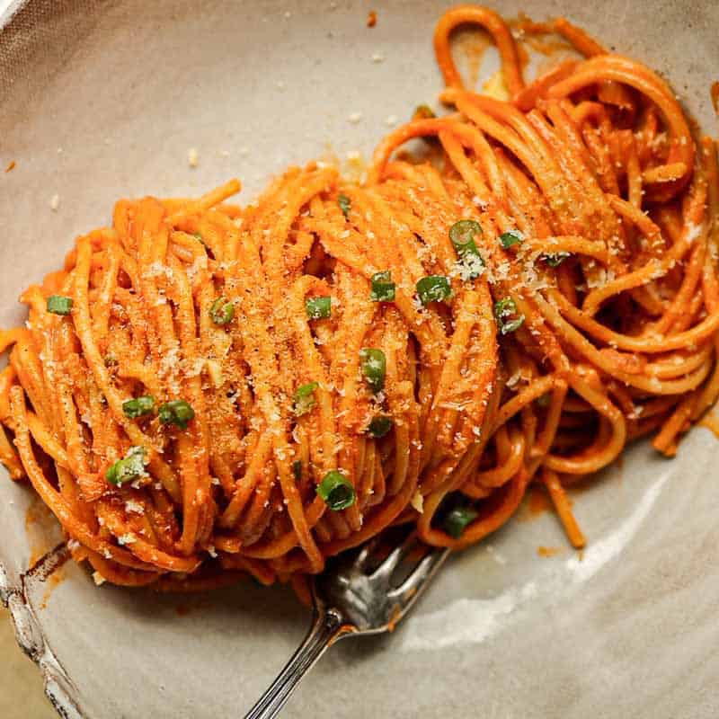 30-Minute Delicious Asparagus Pasta Recipe (Vegan). 21 A close-up of a bowl of vegan gochujang vodka pasta coated in a rich red sauce, garnished with grated cheese and chopped green herbs. A fork rests beside the pasta on the white ceramic plate.
