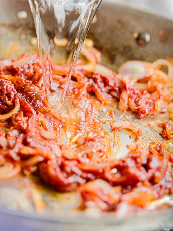 30-Minute Creamy Gochujang Vodka Pasta - Delicious & Dairy-FREE. 17 Close-up of a pan with tomato paste, sliced onions, and spices being mixed, while vodka is being poured in. The ingredients are sizzling, giving an impression of a cooking process.